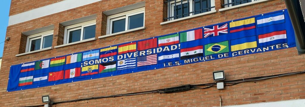 Collage de banderas colgados en la pared sobre fondo azul, con la leyenda "Somos diversidad. IES Miguel de Cervantes".